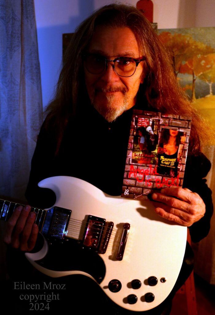 A man holding the Sleepaway Camp book and a white electric guitar.