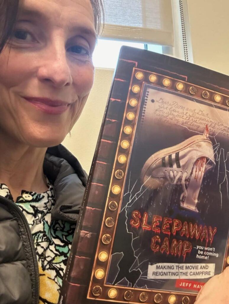 A woman smiles while holding the Sleepaway Camp book beside her face.