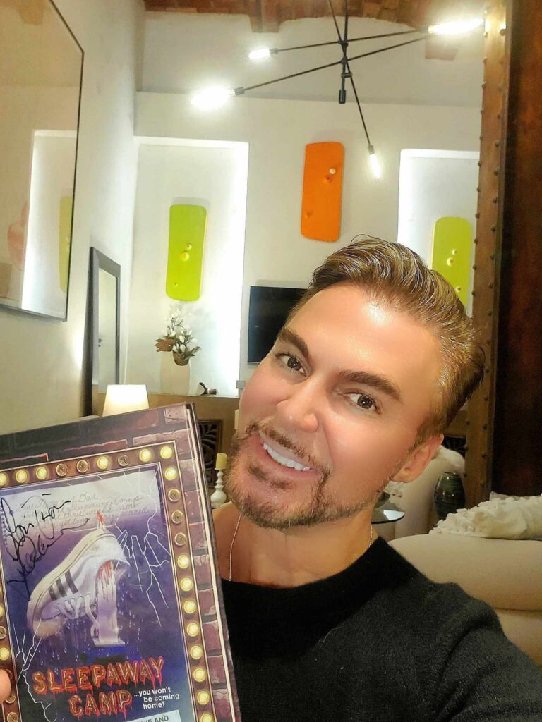A man smiling beside a signed copy of the Sleepaway Camp book.