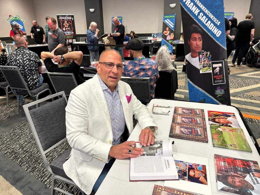 Frank Saladino seated at his convention signing table with an open Sleepaway Camp book and a pen.