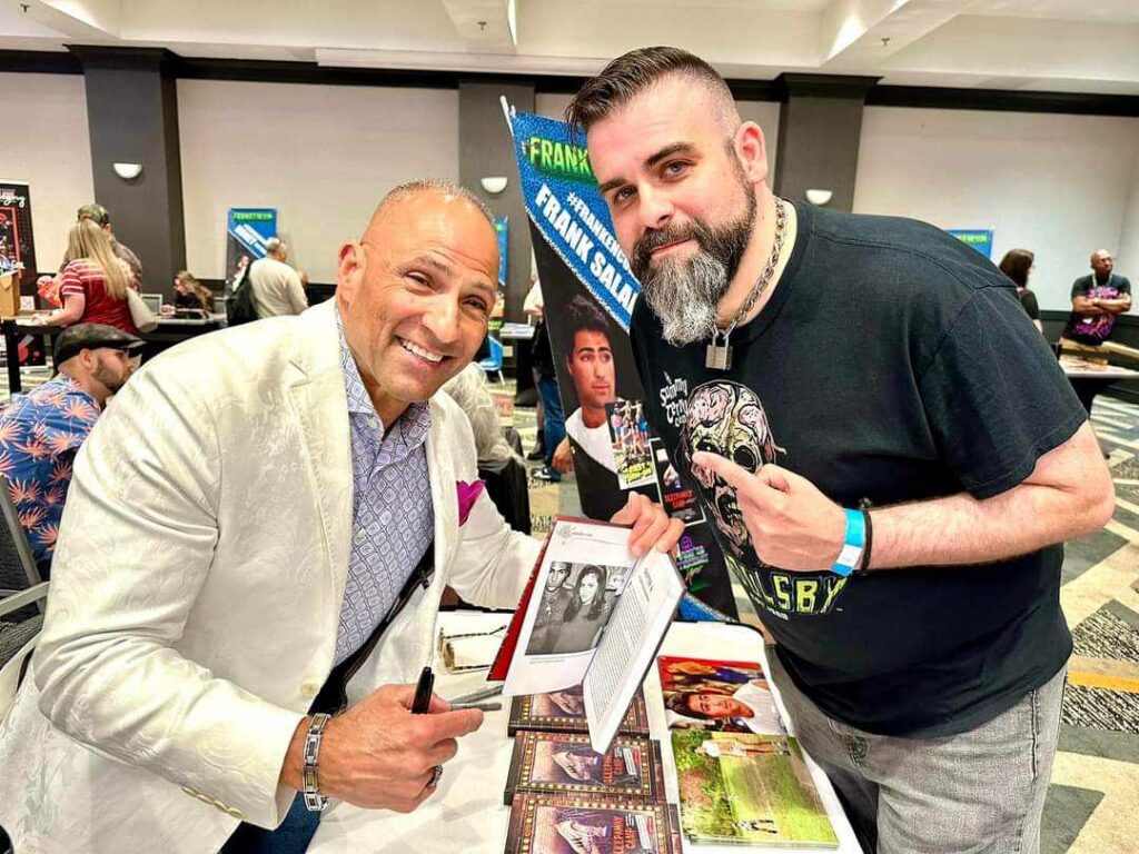 Frank Saladino and another man posing with an open Sleepaway Camp book at a convention signing table.