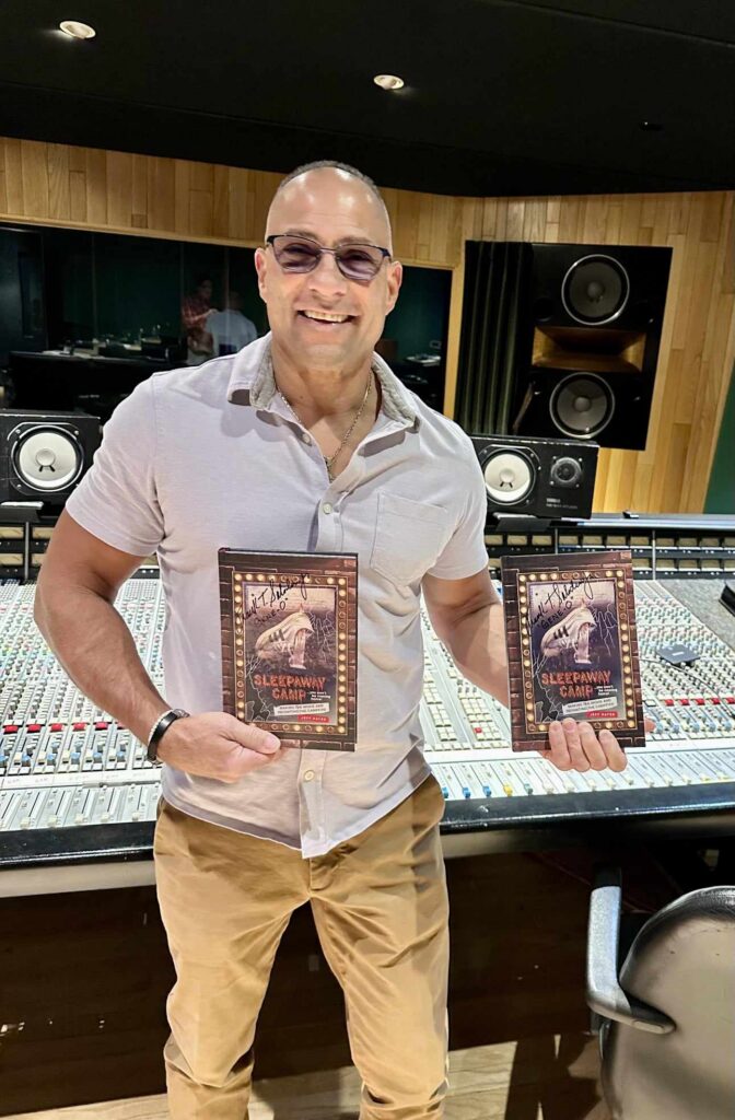 A man holding two copies of the Sleepaway Camp book beside a recording-studio mixing console.