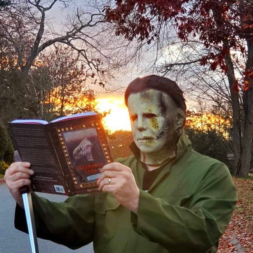 A person wearing a white horror mask reads the Sleepaway Camp book outdoors at sunset.